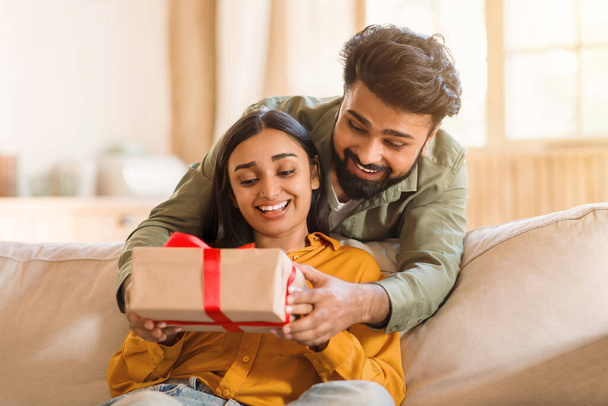 Entzücktes junges indisches Paar drinnen, Frau mit Geschenk in rotes Band gehüllt, teilen Eheleute herzerwärmenden Moment und betonen die Freude am Geben und Empfangen von Überraschungen - Foto, Bild