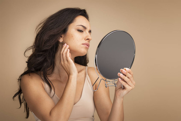 Stressed sad brunette young woman wearing beige top looking at mirror and touching her face over beige studio background, having skin problems. Acne, pimples, wrinkles, dull skin - Zdjęcie, obraz