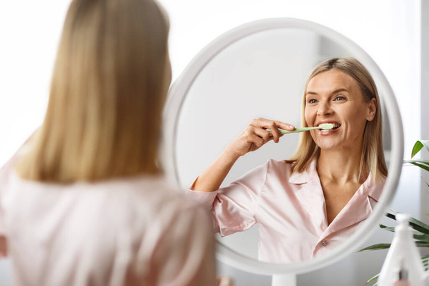 Sabah hijyeni. Aynanın yanında dişlerini fırçalayan çekici olgun bir kadının portresi diş fırçasını tutan ve yansımasına gülümseyen, seçici odaklanan mutlu orta yaşlı kadın. - Fotoğraf, Görsel