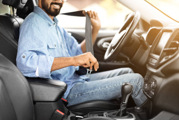 Przycięty przystojnym brodatym młodym Hindusem, noszącym luźny strój kierowcy siedzącego na przednim siedzeniu, zapinającego pasy, jeżdżącego swoim nowym, ładnym samochodem i uśmiechniętym, kopiującym przestrzeń. Koncepcja bezpieczeństwa jazdy - Zdjęcie, obraz