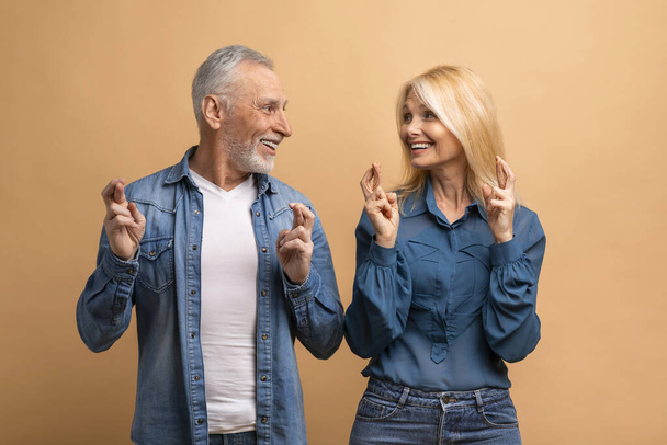 Ευτυχισμένος ηλικιωμένος άνδρας και γυναίκα σταυρώνουν τα δάχτυλα και κοιτάζονται, ενθουσιασμένοι συνταξιούχοι κάνουν μια ευχή, ελπίδα για καλή τύχη, ποζάροντας σε μπεζ φόντο στούντιο. Ανθρώπινες χειρονομίες - Φωτογραφία, εικόνα