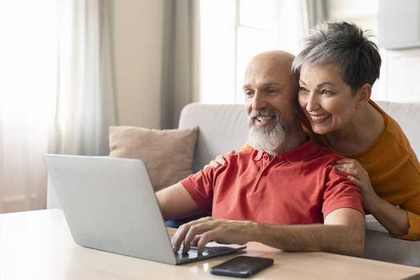 自宅でラップトップを使用して幸せなシニアカップルの肖像画, リビングルームでコンピュータで休む笑顔の高齢者の配偶者, ショッピングオンラインまたは一緒に写真を見る, 背後から夫を抱擁する妻 - 写真・画像