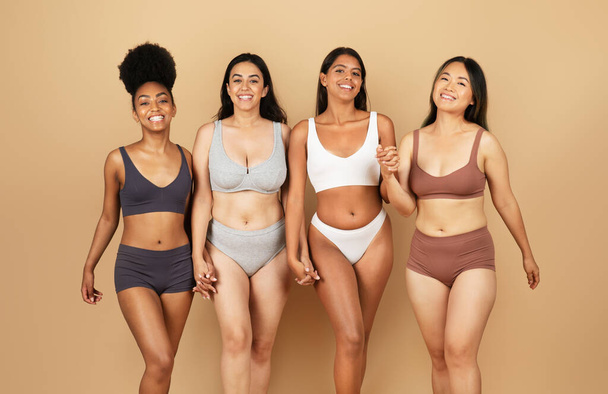 Čtyři různé ženy vyzařují radost a sebedůvěru, stojí spolu v jednoduchém spodním prádle, objímají svou přirozenou krásu na béžovém pozadí - Fotografie, Obrázek