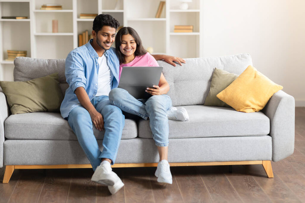 Happy young Indian spouses comfortably seated on sofa, engaged in leisurely browsing or working on laptop computer in homey setting - Photo, Image