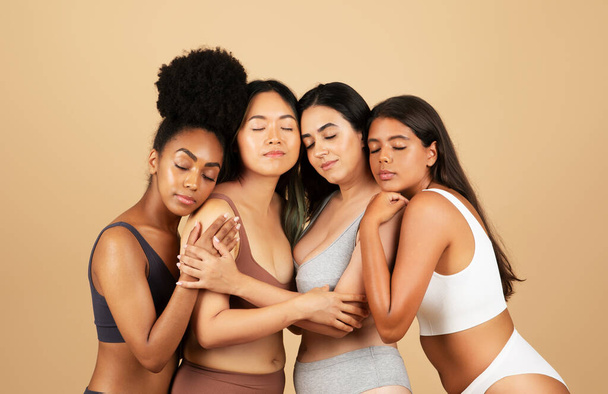 Quattro donne diverse irradiano bellezza naturale mentre si avvicinano, indossando biancheria intima semplice e condividendo un abbraccio confortante sullo sfondo beige neutro - Foto, immagini