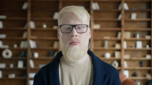 Un serio empresario albino posando ante la cámara con los ojos cerrados. Retrato de un hombre confiado de pie en la biblioteca con estantes de libros en el fondo. Cara de chico pensativo con anteojos, cámara lenta  - Metraje, vídeo