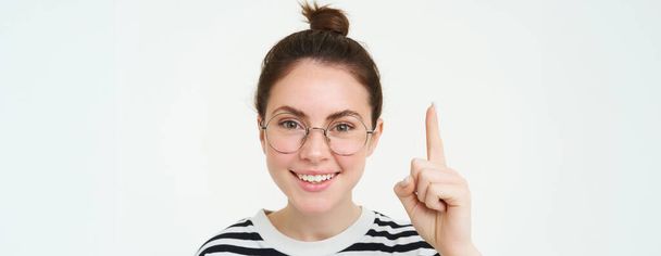 Immagine di donna in occhiali, che punta il dito verso l'alto, mostrando pubblicità in alto, in piedi su sfondo bianco. Copia spazio - Foto, immagini