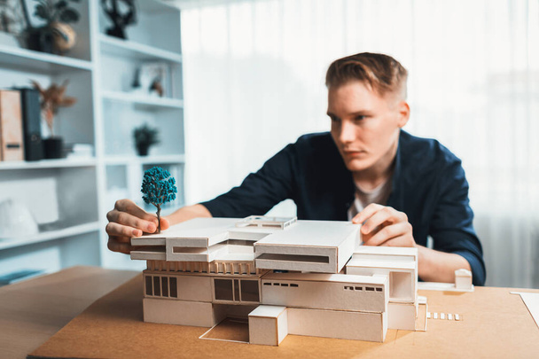 Архітектор-дизайнер вивчає елегантну модель будинку, переглядаючи дизайн споруди для поліпшення з планом будівництва на столі. Творчість та інновації в архітектурному дизайні. Ітерація - Фото, зображення