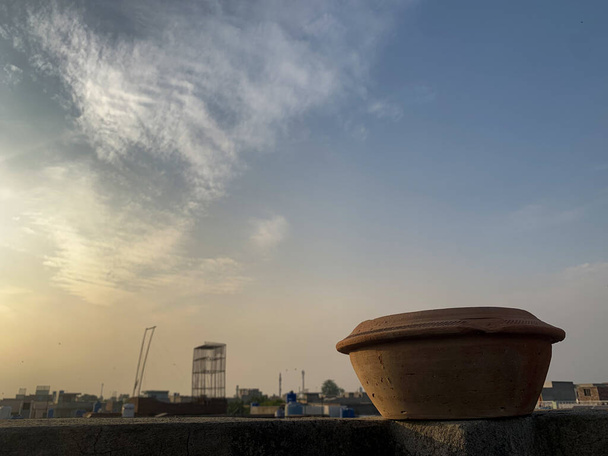 Mud water pot geplaatst op de top van het dak muur. Vogel water feeder gebruiksvoorwerpen met zonlicht en een bewolkte lucht op de achtergrond. - Foto, afbeelding