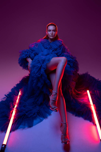 Un modelo impresionante en un lujoso vestido de carnaval azul posando elegantemente por el cálido brillo de las luces de neón en un estudio. - Foto, imagen