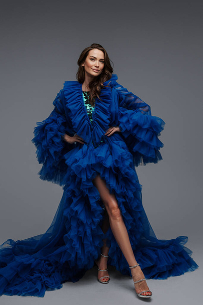 Un modelo impresionante con una sonrisa encantadora en un vestido de carnaval azul exuberante, posando sobre un fondo gris - Foto, Imagen