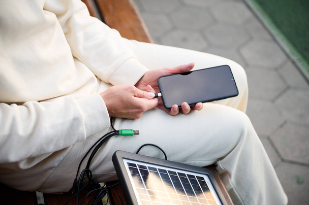 Όμορφη γυναίκα κάθεται στον πάγκο και φορτίζει το τηλέφωνό της με ένα φορητό ηλιακό πάνελ. Ηλιακός φορτιστής μπαταρίας για κινητές συσκευές. Έννοια του βιώσιμου τρόπου ζωής και πράσινης ανανεώσιμης ενέργειας. Προβολή περικοπών - Φωτογραφία, εικόνα