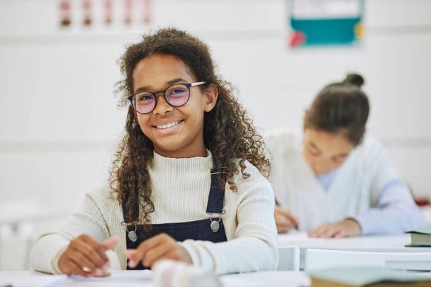 Muotokuva hymyilevä musta koulutyttö silmälasit ja katsot kameraa istuessasi työpöydän ääressä koulussa, kopioi tilaa - Valokuva, kuva