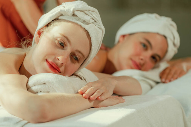 Ein paar schöne junge Frauen mit weißem Handtuch liegen auf einem bequemen Wellness-Bett während einer entspannenden Körpermassage durch einen professionellen Masseur im Wellness-Salon umgeben. Grauer Hintergrund. Gelassenheit. - Foto, Bild