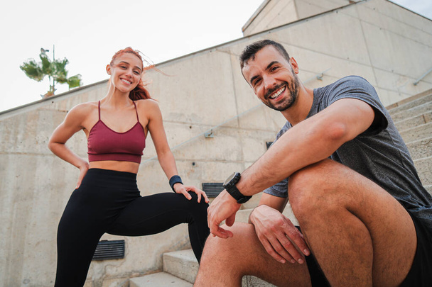 Smiling sporty couple resting after a workout, enjoying a healthy lifestyle after training together. Young male and female runners dressed in sportswear looking at camera. Athletic personal trainer - Фото, зображення
