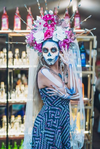 Мексиканка, одетая и охарактеризованная как катрина ко Дню Мертвых, несет в руках бутылку мезокала со скорпионом внутри, позади нее фон из бутылок мезокала. - Фото, изображение
