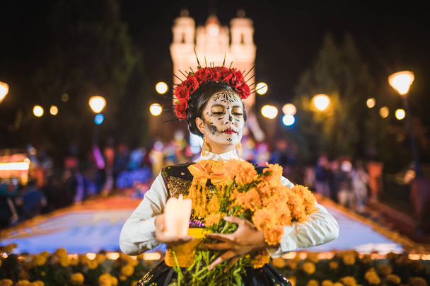 Мексиканская женщина, одетая и охарактеризованная как катрина ко Дню Мертвого Праздника, с цемпасучильными цветами и свечами, за ней подношение мертвых. - Фото, изображение