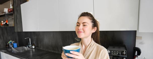 Retrato de mujer sonriente bebe café, se para en la cocina y disfruta de deliciosa taza de capuchino por la mañana, se ve feliz ante la cámara. - Foto, Imagen