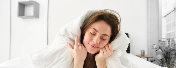 Retrato de una chica con sueño después de levantarse temprano, acostada en la cama debajo de una manta y sonriendo. - Foto, Imagen