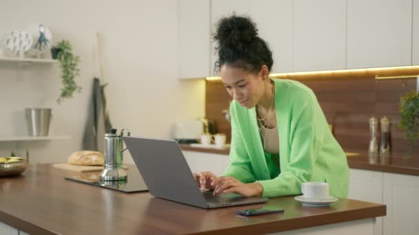 Vielrassige junge Frau in lässiger Kleidung steht in der modernen Küche, lehnt am Esstisch, schaut auf Laptop und tippt, wählt Lebensmittelgeschäft im Online-Shop, macht Essenslieferung im Internet - Filmmaterial, Video