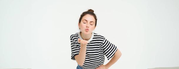Portrait of beautiful brunette woman sends air kiss at camera, holds palm near lips, isolated against white background. - Photo, Image