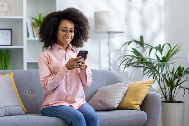 Jonge mooie vrouw zit op de bank in de woonkamer van het huis thuis, Afro-Amerikaanse vrouw glimlachen en het houden van telefoon, het typen van bericht met behulp van smartphone app, glimlachen en gelukkig. - Foto, afbeelding