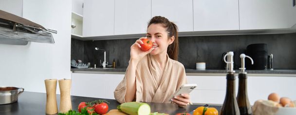 Portret młodej kobiety zamawiającej zakupy na smartfonie, trzymającej pomidory, siedzącej przy desce z warzywami. Dziewczyna szuka przepisu online, za pomocą telefonu komórkowego. - Zdjęcie, obraz