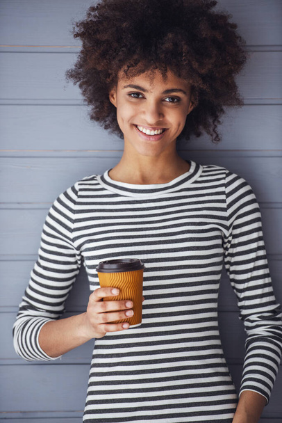 Stylowa dziewczyna trzyma filiżankę kawy, patrząc w aparat i uśmiechając się, na szarym tle - Zdjęcie, obraz