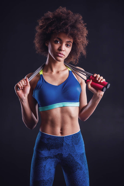 Attractive girl in sportswear is holding a skipping rope and looking at camera, on black background - Photo, Image