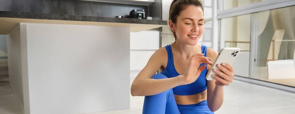 Imagem de jovem mulher em forma vestindo sportbra e leggings azuis, usando o aplicativo smartphone, faz exercício em casa, senta-se no chão e segue vídeo de treinamento on-line. - Foto, Imagem