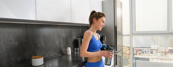 Εικόνα του νεαρού γυμναστή, γυναίκα σε αθλήματα και κολάν, κρατώντας το μπουκάλι του νερού, πίνοντας μετά την προπόνηση, στέκεται στην κουζίνα. - Φωτογραφία, εικόνα