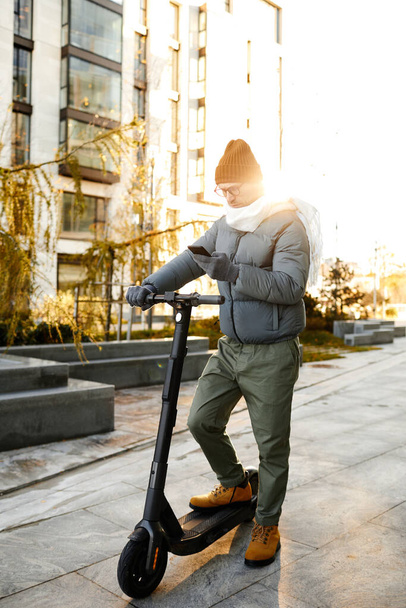 Joven con ropa de abrigo conectando scooter con su teléfono móvil para pasear por la calle - Foto, Imagen