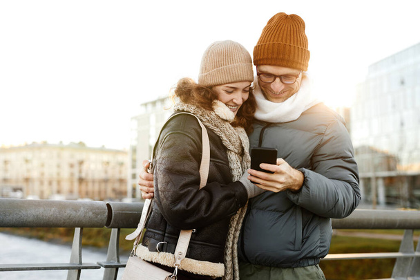Молодая счастливая пара смотрит фотографии на мобильном телефоне, стоящем на улице - Фото, изображение