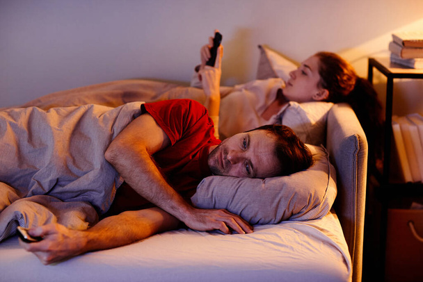 Young man lying on bed and watching TV with his girlfriend using mobile phone before sleep in background - Fotó, kép