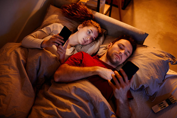 Vista de alto ângulo do jovem casal usando gadgets antes de dormir enquanto deitado na cama no quarto - Foto, Imagem