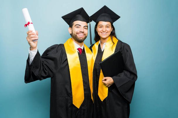 Opgewonden gelukkige jonge man en Spaanse vrouw met afstuderen jurk afwerking van de universiteit samen en tonen hun universitaire diploma - Foto, afbeelding