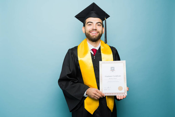 Mexikanischer smarter junger Mann lächelt und zeigt stolz sein Universitätsdiplom mit seinem Abschlusskleid vor blauem Studiohintergrund - Foto, Bild