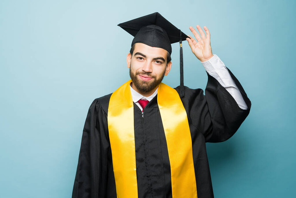 Gut gelaunter, attraktiver junger Mann, der sich Mütze und Kleid aufsetzt, um nach seinem Universitätsabschluss sein College-Diplom zu erhalten - Foto, Bild