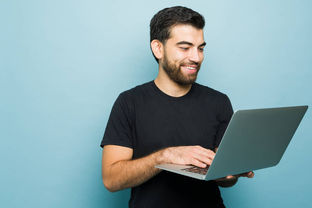 Szczęśliwy młody człowiek i student uśmiechnięty za pomocą laptopa podczas odrabiania lekcji online lub robi zakupy online na niebieskim tle - Zdjęcie, obraz