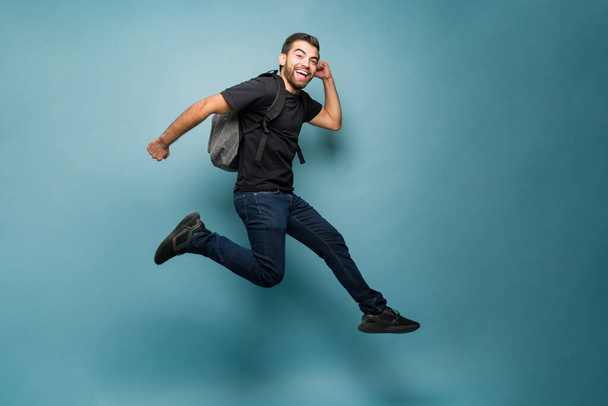 Ενθουσιασμένος φοιτητής που δείχνει χαρούμενος πηδώντας στον αέρα διασκεδάζοντας ενώ πηγαίνει στο πανεπιστήμιο με φόντο ένα μπλε στούντιο - Φωτογραφία, εικόνα