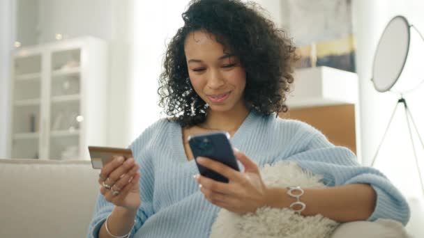 Χαμογελώντας πολυφυλετική γυναίκα κρατώντας smartphone και πιστωτική κάρτα κάθεται στον καναπέ σύγχρονο σπίτι. Ευτυχής ποικιλόμορφη γυναίκα αγοραστής χρησιμοποιώντας άμεσες εύκολες πληρωμές για κινητά, πελάτης κάνοντας την αγορά online τραπεζική εφαρμογή - Πλάνα, βίντεο