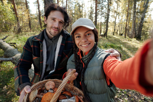 Young intercultural couple of mushroom pickers taking picture on mobile phone while both looking at camera in autumn forest - Photo, Image