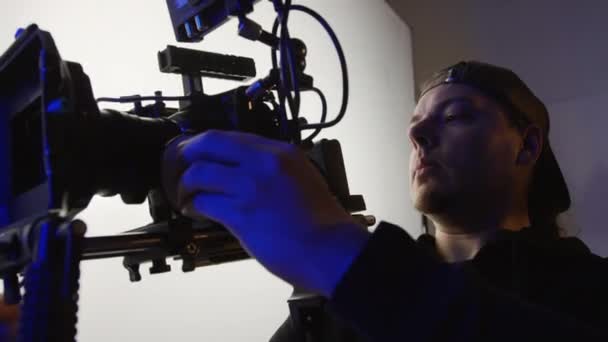 Tiro médio de cinegrafista caucasiano em boné de beisebol segurando câmera de vídeo profissional na plataforma de ombro, ajustando quadro e conversando com a equipe de produção, enquanto trabalhava nos bastidores em filmagens comerciais - Filmagem, Vídeo