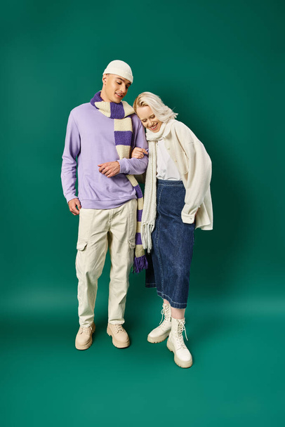 стильна пара в зимовому вбранні, щаслива жінка позує з чоловіком в теплому одязі на бірюзовому тлі - Фото, зображення