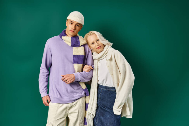 молодая пара в зимнем наряде, счастливая женщина позирует с мужчиной в стильной одежде на бирюзовом фоне - Фото, изображение