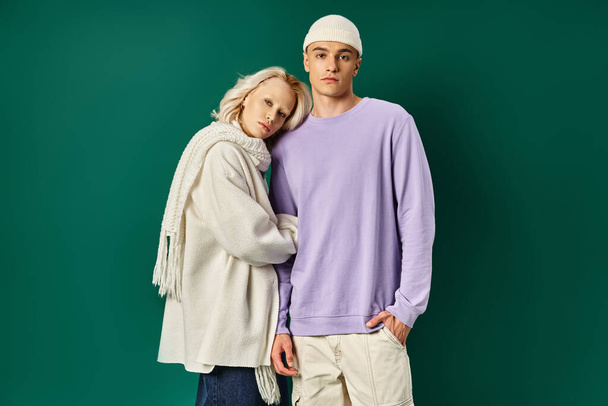 tall handsome man in beanie hat posing with blonde woman in winter attire on turquoise backdrop - Photo, Image