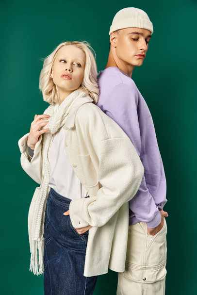 blonde woman in winter attire posing with hand in pocket and back to back with tall man on turquoise - Photo, Image