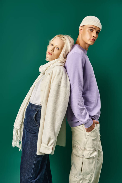 blonde woman in winter attire standing back to back with young man in beanie on turquoise backdrop - Photo, Image
