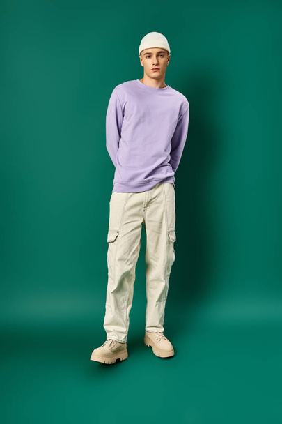 full length of man in purple sweatshirt and beanie hat posing on turquoise backdrop, winter fashion - Foto, immagini