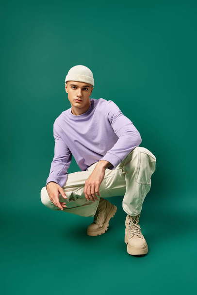 full length of man in purple sweatshirt and beanie hat sitting on turquoise backdrop, winter fashion - Foto, immagini